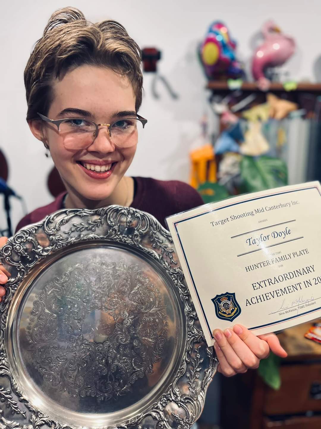 Taylor is smiling as she holds up a pewter award plate and certificate that she won for target shooting.