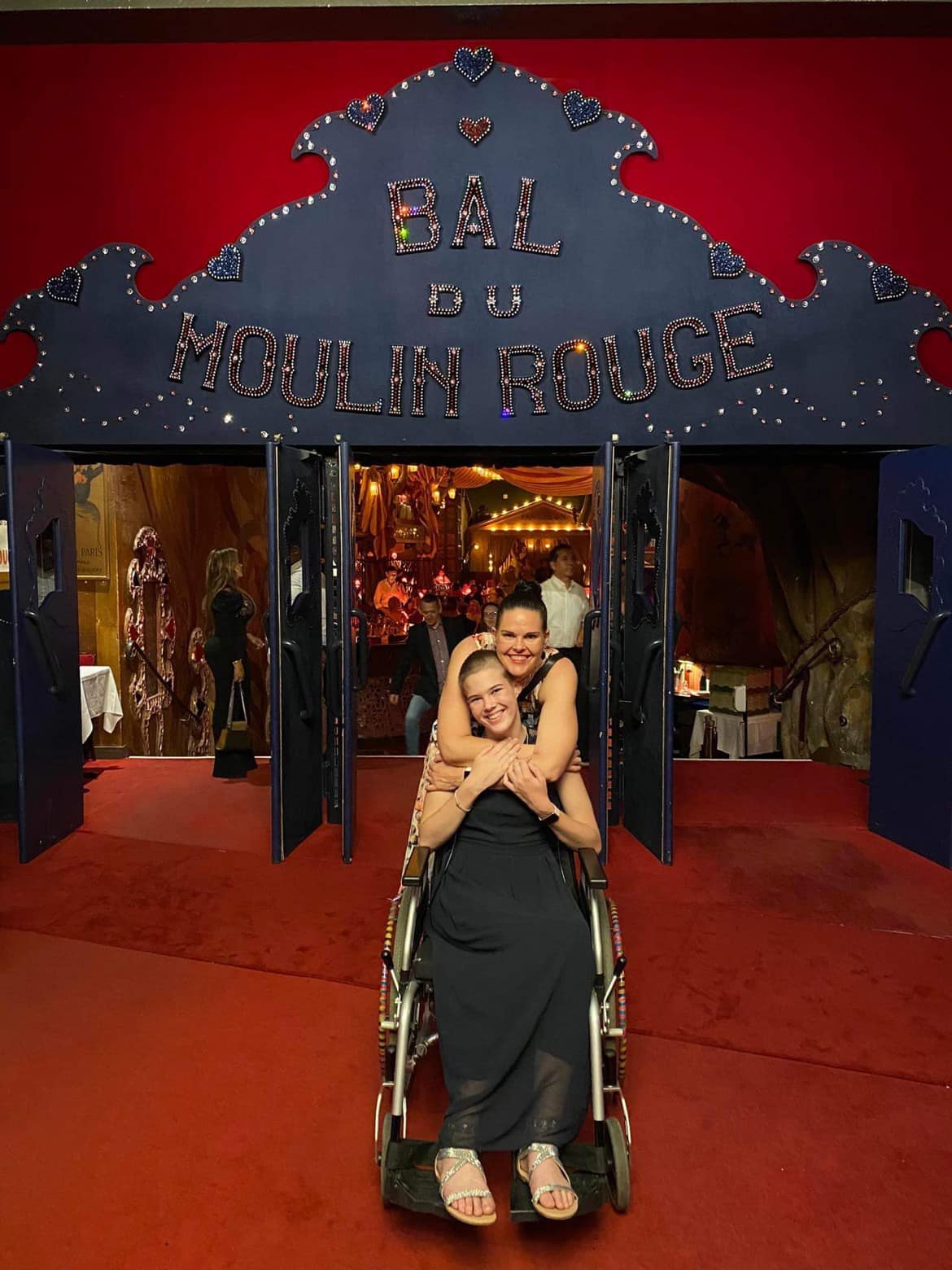 Taylor is sitting in a wheelchair in front of the entrance to the Moulin Rouge. Her mother Zalie is embracing her from behind and they are both smiling at the camera.