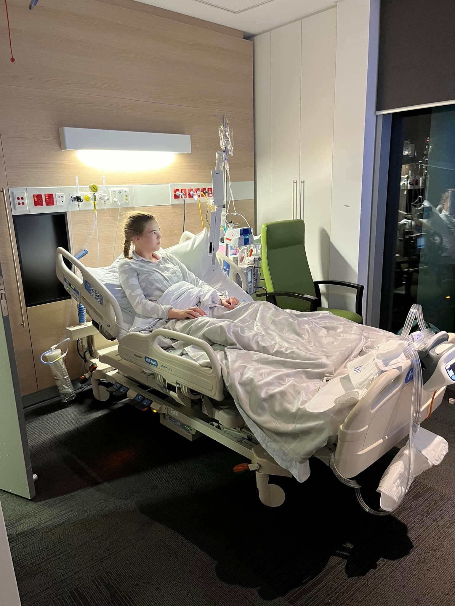Photograph of a teenage girl in a hospital bed, she is sitting up and looking left out of the window.